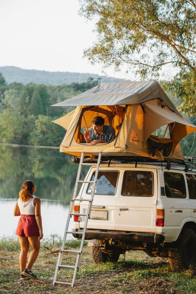 Back view of unrecognizable young female traveler standing on lake shore and talking to boyfriend resting in tent placed on SUV car roof